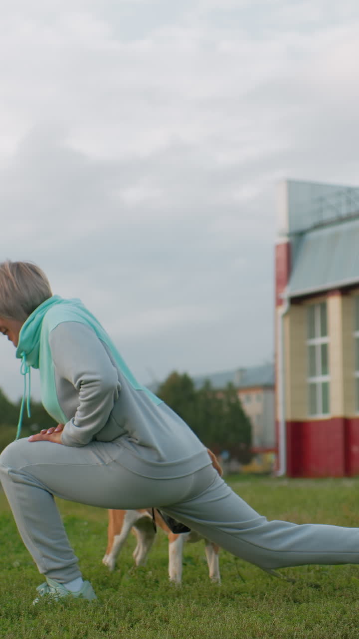 Mujer mayor estirándose con perro beagle, suave zancada en un campo urbano cerca de un edificio escolar, correa atada al collar, cielo nublado, chándal y bufanda informales, rutina de bienestar matutina