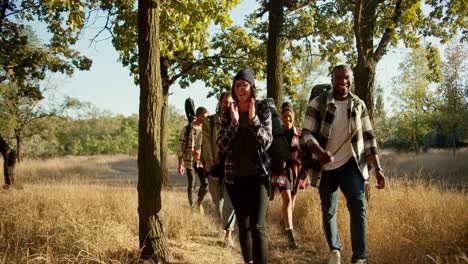 Front-view-of-a-happy-group-of-several-pairs-of-people-in-special-hiking-clothes-with-hiking-backpacks-walking-through-the-forest-near-dried-yellow-summer-grass.-A-Black-man-talks-to-a-blonde-girl-with-a-bob-hairstyle