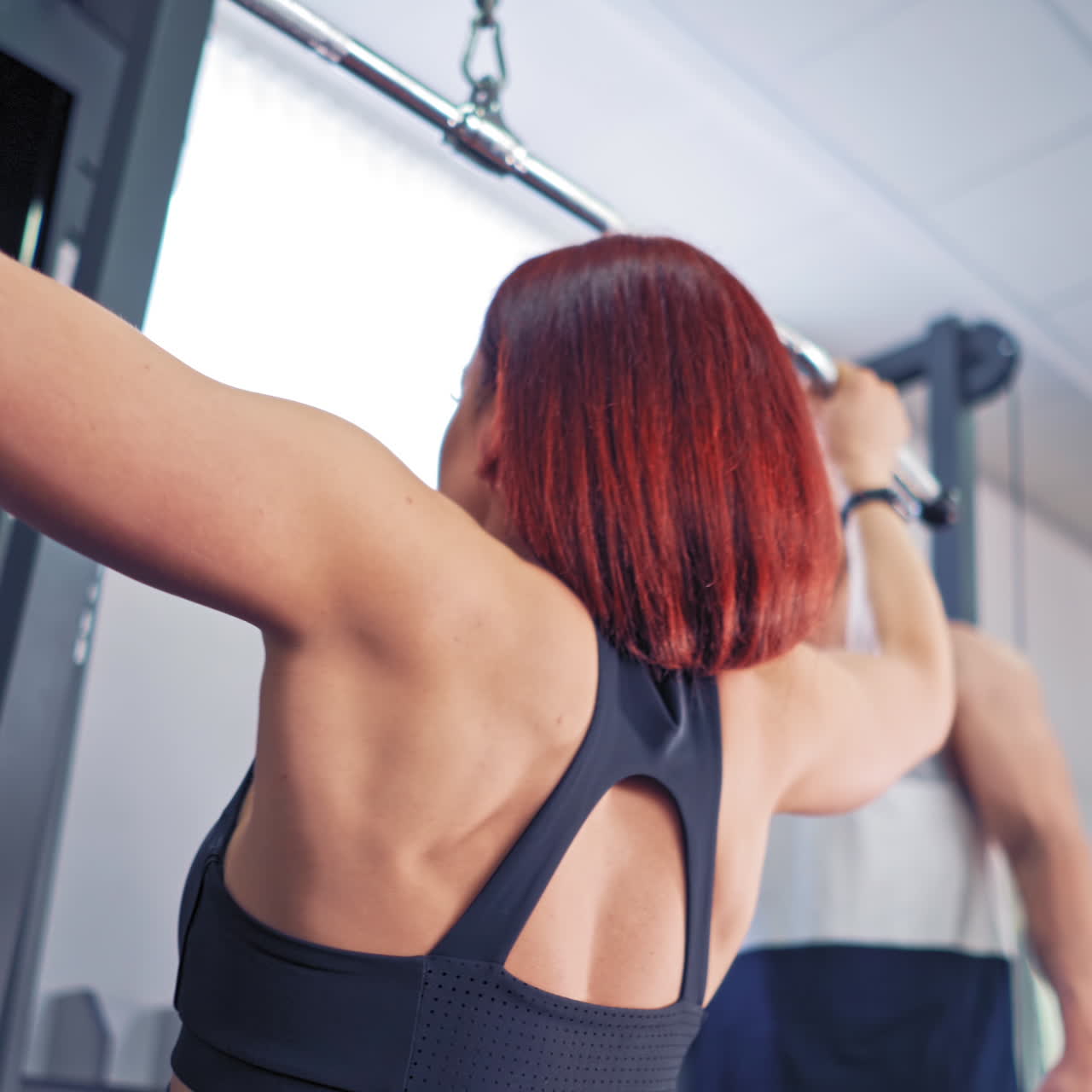 Back view of strong woman exercising on a modern simulator. Red-haired sportswoman pulling the bar down and her instructor controls the process in the gym.