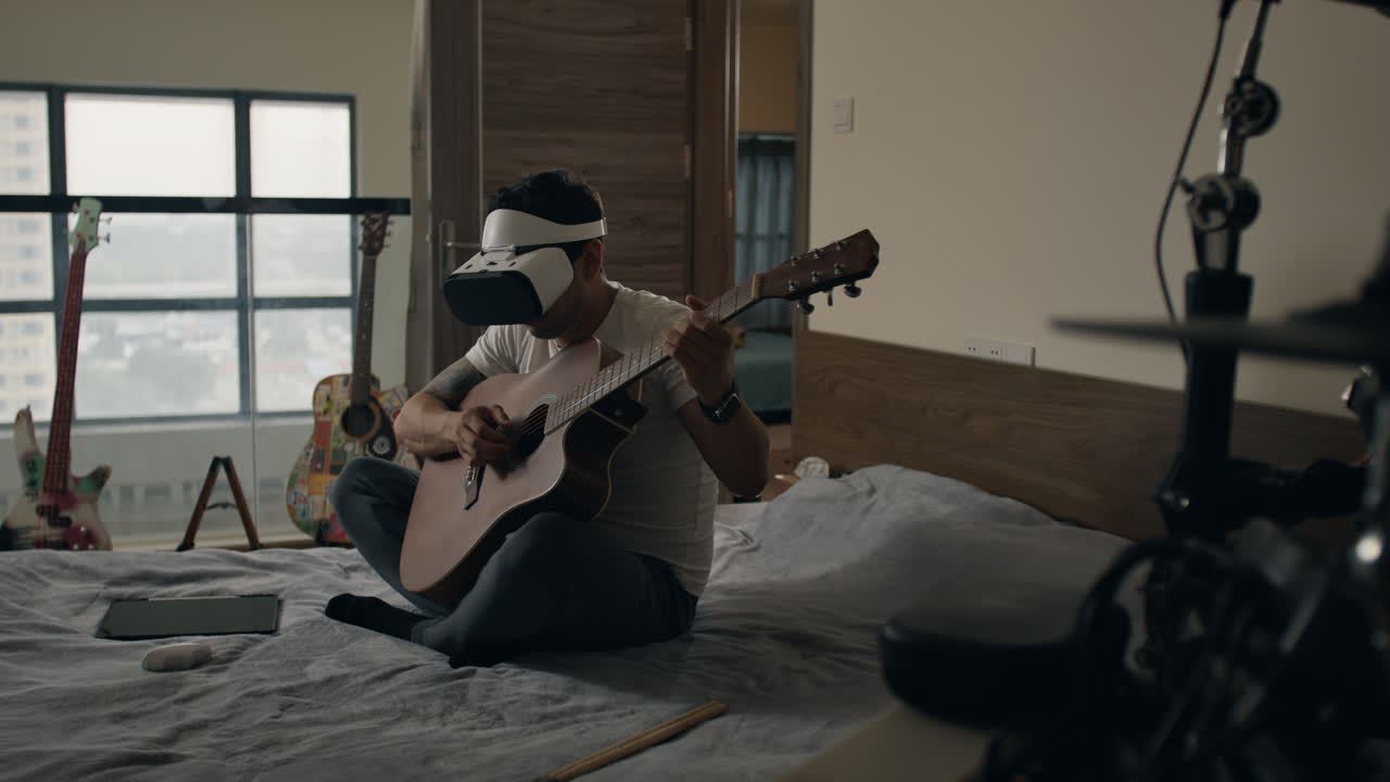 Man wearing VR headset plays acoustic guitar while sitting on bed in apartment bedroom
