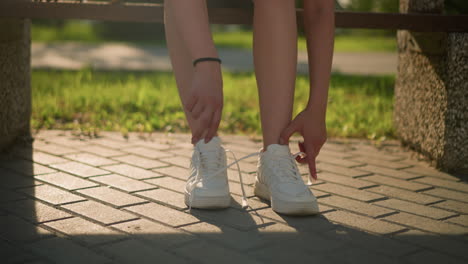 leg view of lady loosening shoelace of her left sneaker while sitting on bench outdoors, greenery in background, bright sunlight creates soft shadows on pavement