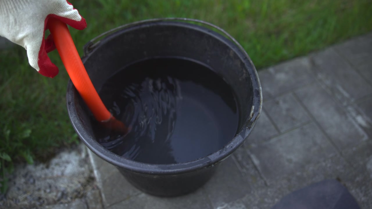 Topbillede af en sort plastikspand fyldt med vand, der står på en betonsti ved siden af grønt græs i blødt dagslys, i et simpelt udendørs miljø.