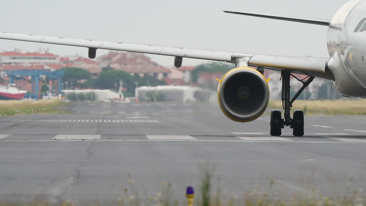 Close-up view of airplane wing and engine on runway, showcasing aircraft details and airport facilities in the background, shot in slow motion