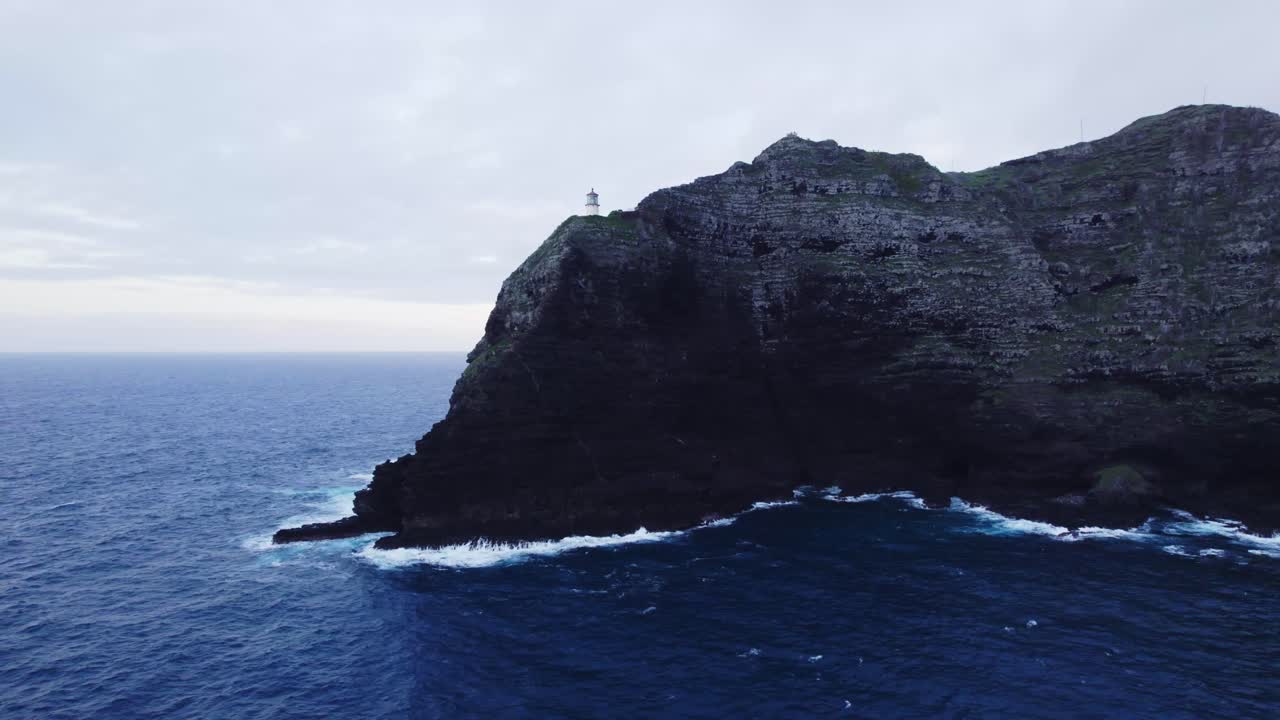 A dramatic aerial shot of Makapu'u Point Lighthouse perched atop a rugged cliff, with the deep blue ocean waves crashing below