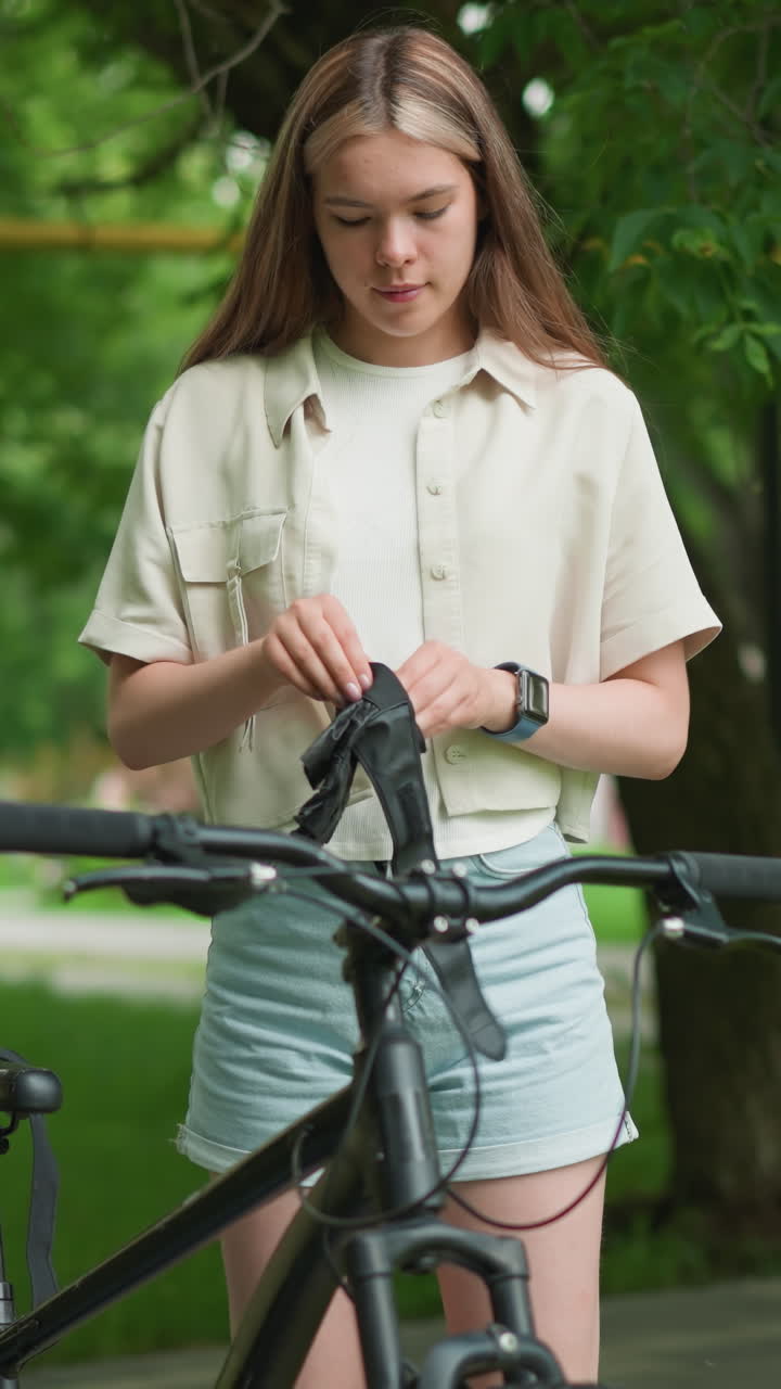 joven adulto ajusta guantes negros al lado de su bicicleta en un camino pavimentado rodeado de exuberante vegetación, lleva un reloj inteligente en la mano derecha mientras el fondo muestra una vista borrosa de los edificios