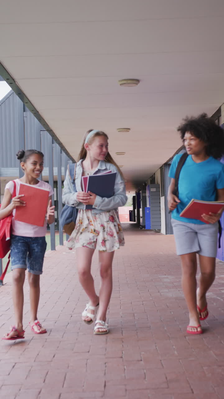Vertical video three diverse, happy schoolgirls talking, walking in school corridor, copy space