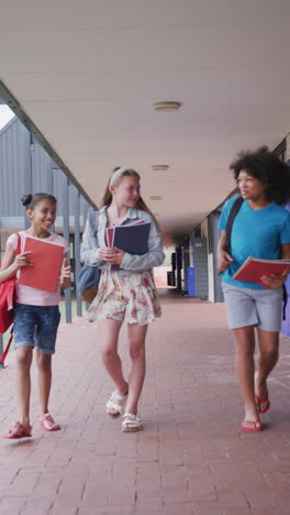 vertical video three diverse, happy schoolgirls talking, walking in school corridor, copy space