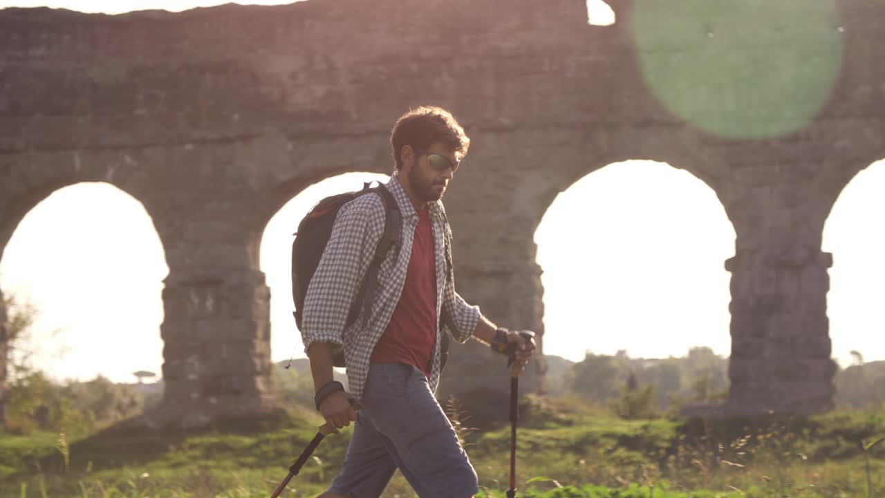 Giovane backpacker cammina a piedi nudi con bastoni sull'erba davanti agli archi dell'acquedotto romano nelle rovine del Parco degli Acquedotti a Roma al tramonto, al rallentatore