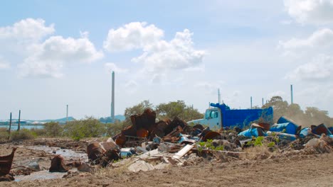 barrels-with-crude-oil-and-water-tanks-lying-on-the-ground-with-a-moving-blue-truck-in-the-background