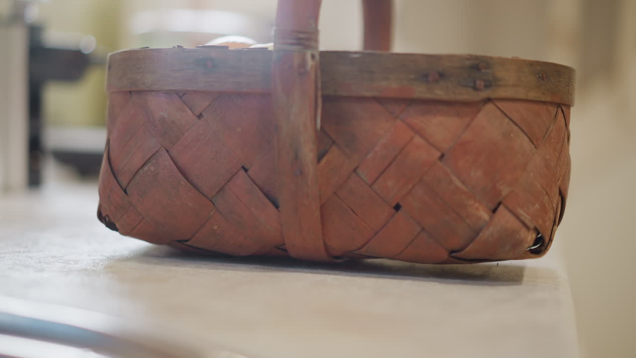 Primo piano di un cesto intrecciato sul bancone della cucina. Un vecchio cesto intrecciato poggia su un piano di lavoro in pietra con una messa a fuoco morbida e poco profonda, luce naturale del mattino e piante e utensili sfocati sullo sfondo, dettaglio di natura morta.