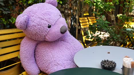 purple teddy bear sitting in an outdoor cafe