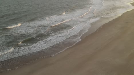 Toma-Aérea-De-Pájaros-De-Una-Persona-Solitaria-Disfrutando-Del-Clima-Caminando-En-La-Playa-De-Arena-Y-Alcanzando-Las-Olas-Del-Océano-Al-Atardecer