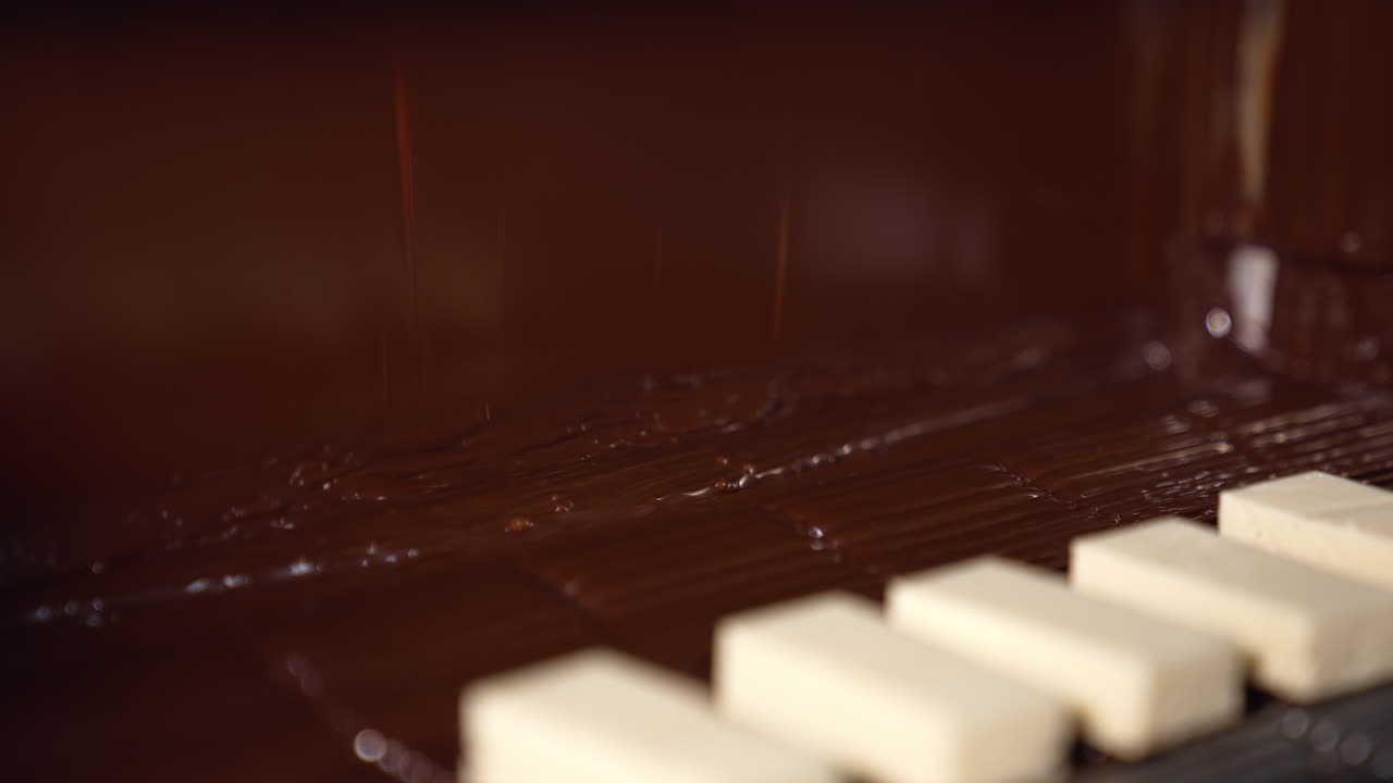 Bars of milky sweets being deepened into a thick layer of black chocolate. Delicious flow of chocolate falling onto the desserts on the conveyor line. Chocolate candies production close up.