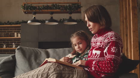 encantadora madre sentada en el sofá con una adorable hija y leyendo su cuento de hadas el día de navidad en casa 1