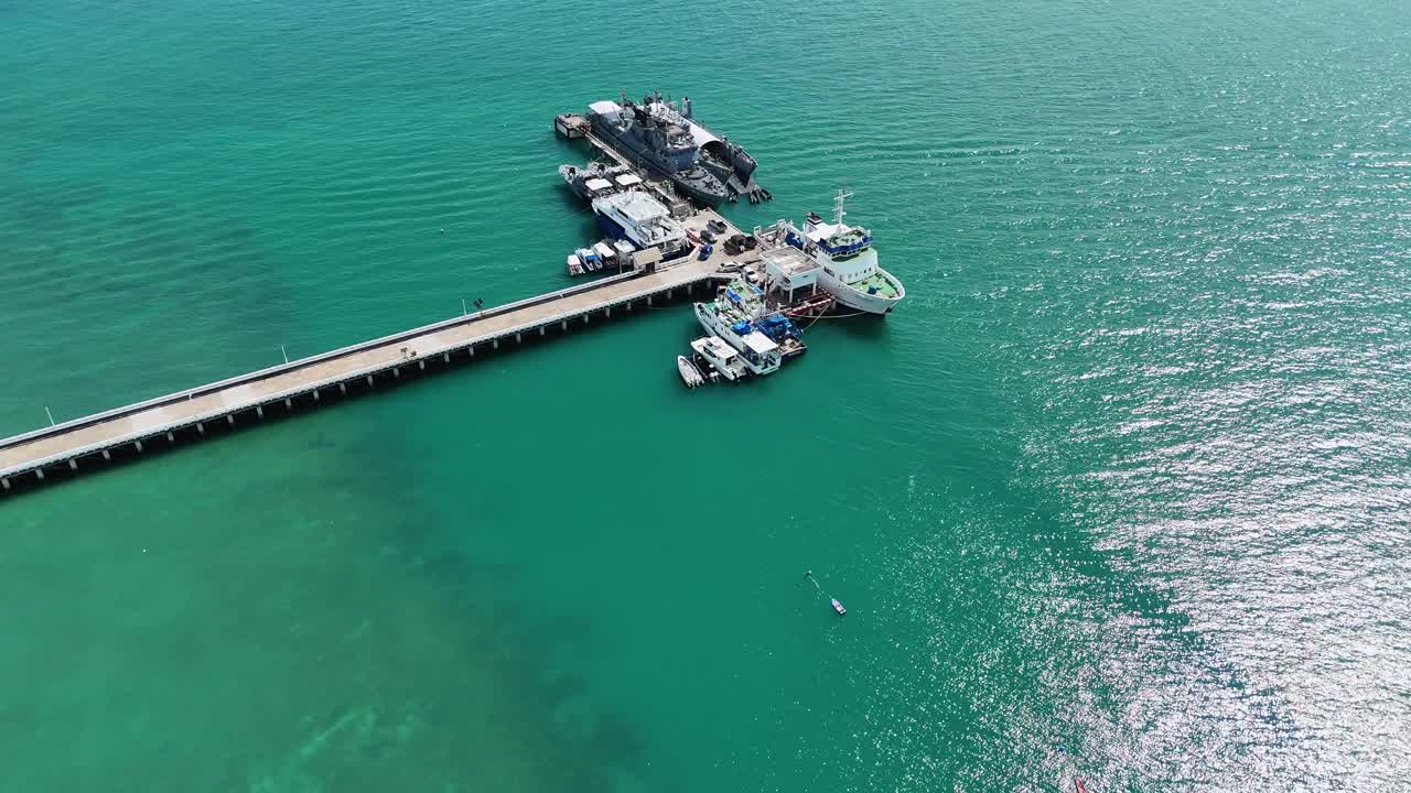 Drone footage captures a bustling pier in Phuket, Thailand, with boats docked in turquoise waters under bright sunlight
