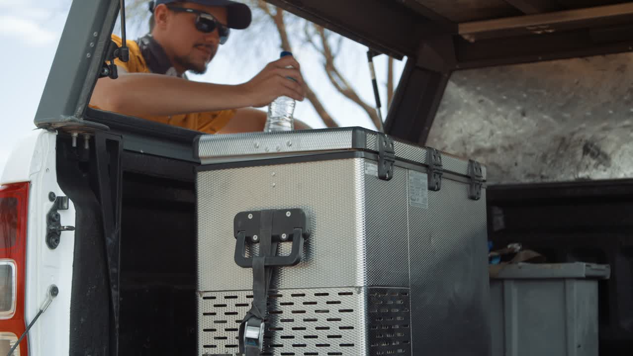 Static shot of a Caucasian male tourist in Africa as he walks towards the back of his rental off-road vehicle and take out a bottle of water from camping vehicle fridge and proceeds to walk away.