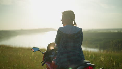 a woman sits alone on a bike in a grassy field, surrounded by open nature with a serene water body in the distance