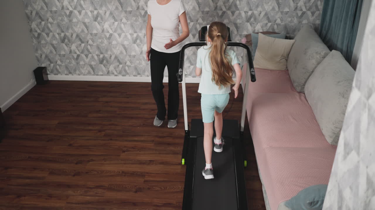 Woman observes daughter walking on treadmill during workout session in cozy home interior featuring patterned wall, soft couch, and wooden floor