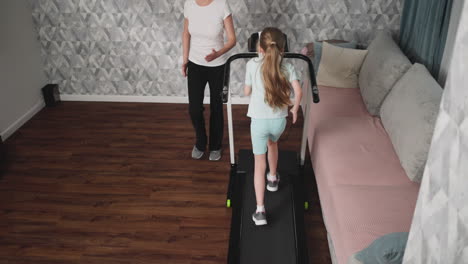 woman observes daughter walking on treadmill during workout session in cozy home interior featuring patterned wall, soft couch, and wooden floor