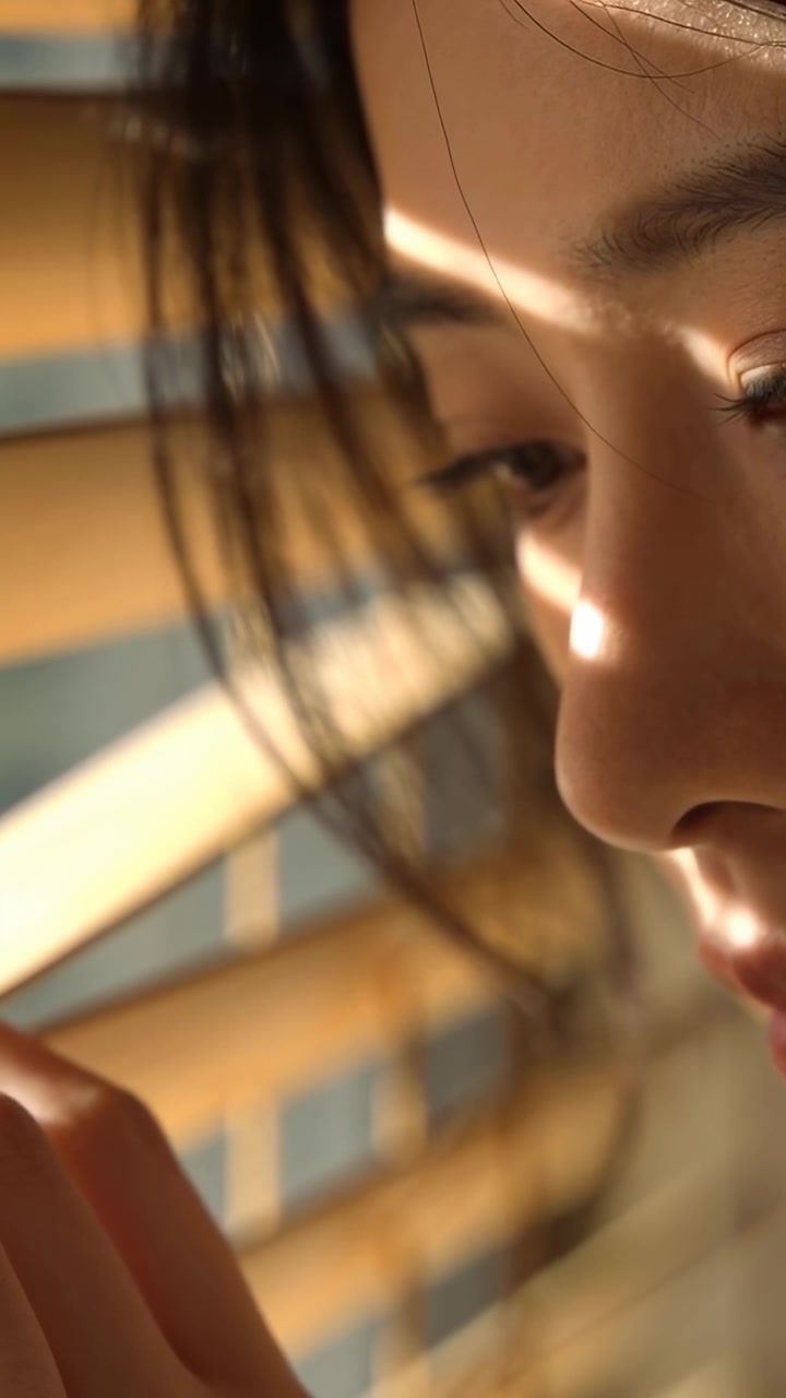 Vertical video: Peering woman holding hand near blinds by window as sun stripe crossing face
