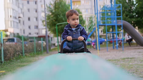 cute toddler sitting on one end of the seesaw. baby boy in jeans jacket on the playground.