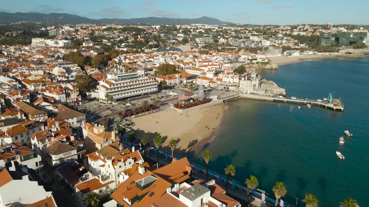 Aerial drone video flying over a calm and luxury beautiful Cascais city shoreline beach and neighbourhood during a sunny summer day with sandy beach, palm trees and red rooftops of white houses seen