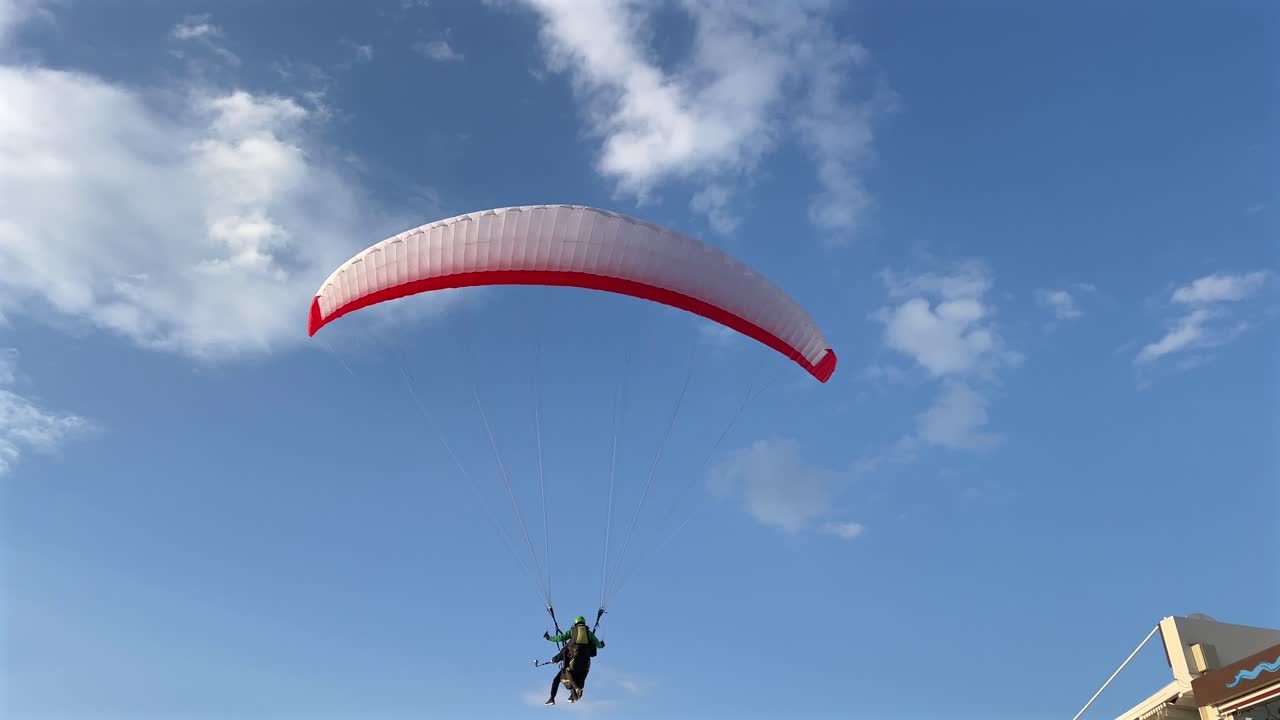 Turkey,Fethiye,Oludeniz,A red and white paragliding glides slowly on the blue skies with white clouds, lands slowly to the lively board walk with people around and in front of the shops by the beach.