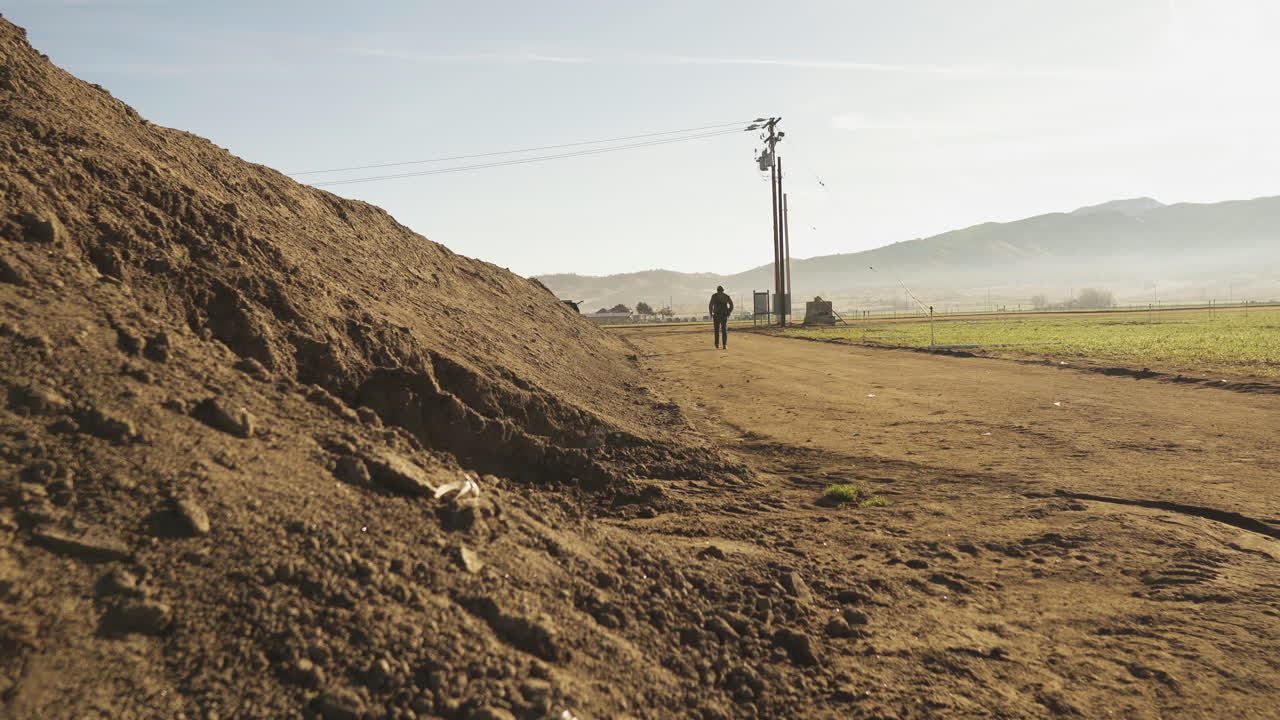 plano general de un hombre caminando por un camino de tierra junto a una berma de tierra y un campo agrícola, con un poste de teléfono y montañas en el fondo