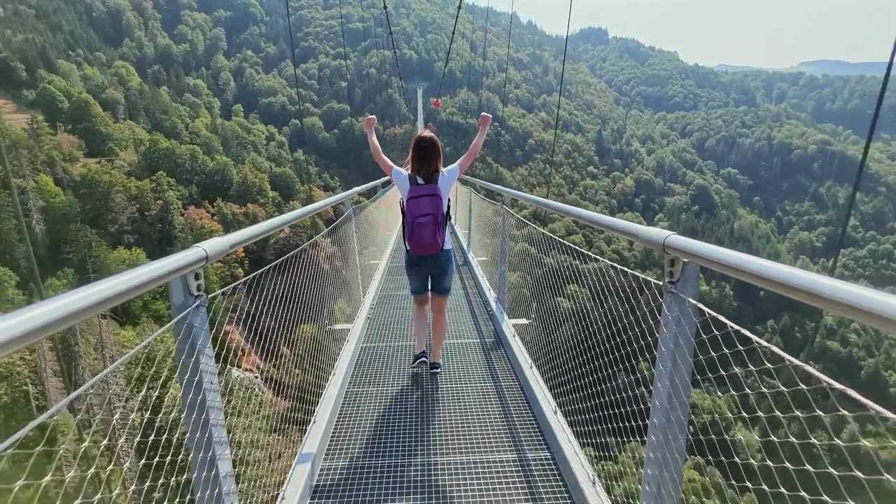 Escursionista donna con uno zaino che si gode la vista panoramica e alza le braccia in segno di vittoria su un lungo ponte sospeso sopra una valle verde