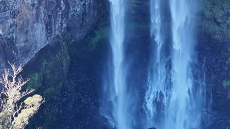 drone camera smoothly descends alongside ebor waterfall, capturing cascading water, rocky cliffs, and lush greenery in bright natural daylight with steady movement