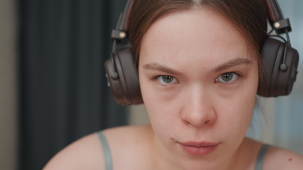 Nærbilde av hvit kvinne med hodetelefoner, intenst fokusert uttrykk, direkte øyekontakt, innendørs studio med mørk gardinbakgrunn, diskrete fregner og naturlig hudtekstur, podkaster eller gamer