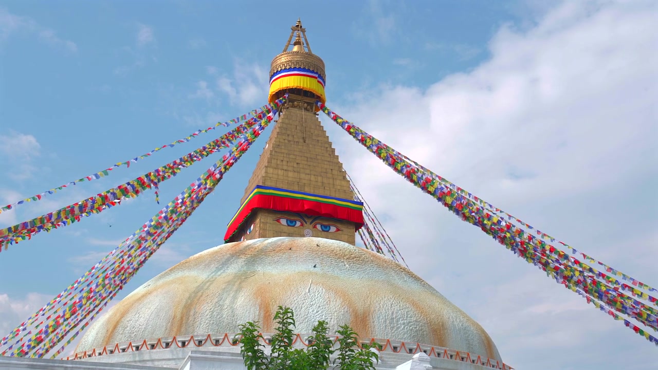 Panoroitu matalan kuvakulman keskikuva, joka keskittyy Boudhanath Stupan kultaiseen torniin ja viisauden silmiin, värikkäiden rukouslippujen ympäröimänä kirkkaan sinisen taivaan alla Nepalissa