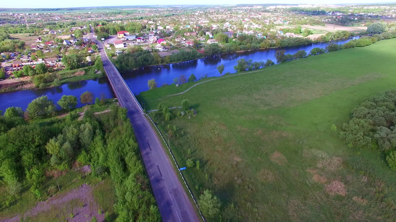 Blue river separating the village from farmlands. Little concrete bridge over the river. Sunny town panorama. Aerial view.