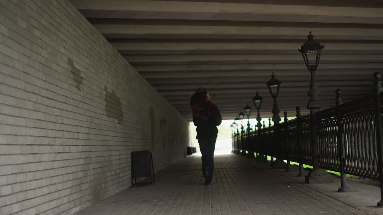 Donna che cammina attraverso un sottopassaggio piovoso, silhouette. Capelli rossi visibili, cappotto e stivali che schizzano pozzanghere, ringhiera in ferro e colonne di lampioni, luce cittadina fioca e nuvolosa, pavimentazione bagnata riflettente, passo costante.