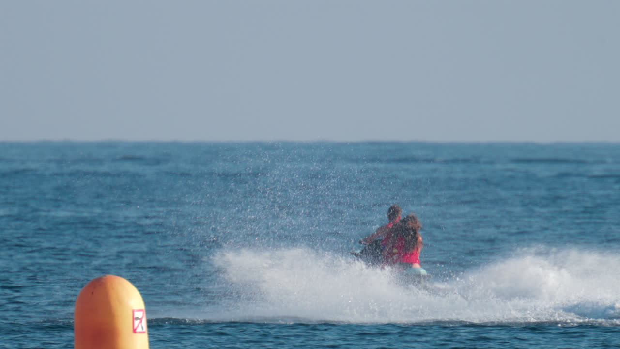 A couple wearing red life vests rides a jet ski across the bright blue sea, splashing water behind them