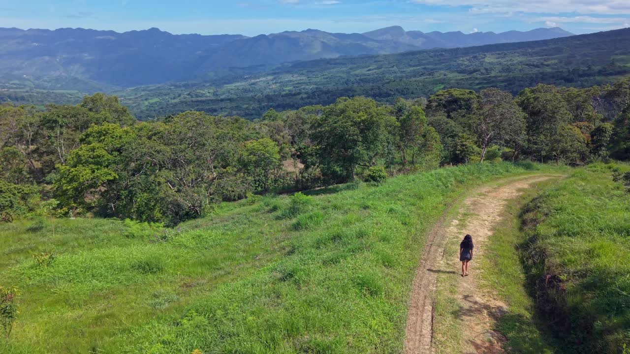 A lone woman walks along a grassy path as mountains and trees unfold beneath a bright sky. A peaceful, poetic aerial capturing freedom, solitude, and connection with nature