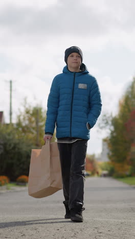 niño ruso con chaqueta azul y gorra negra camina pensativo en el área residencial, una mano en el bolsillo, la otra sosteniendo la bolsa de compras, en medio de vibrantes colores de otoño