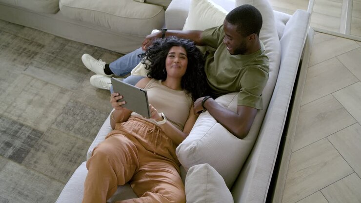 Donna indiana e uomo afroamericano si rilassano su un divano componibile, un tablet scatena risate condivise.