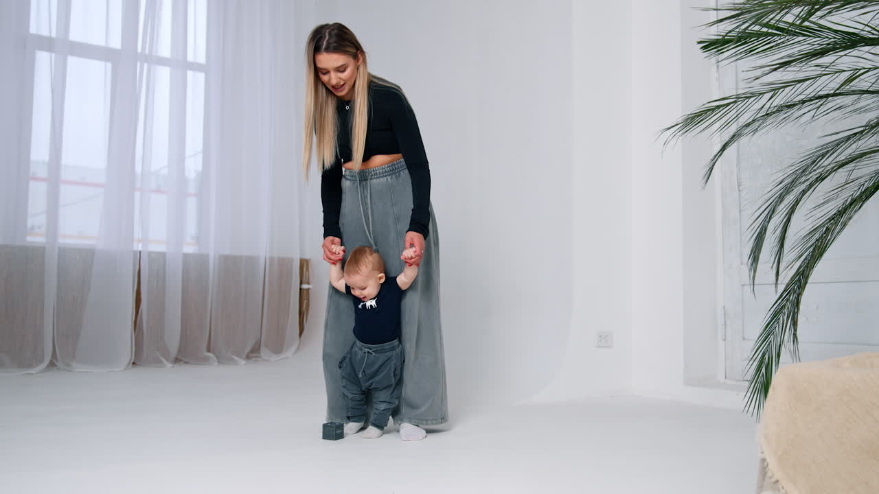 Mother teaching her baby to walk. Long-haired woman holds an infant by the hands. Room backdrop.