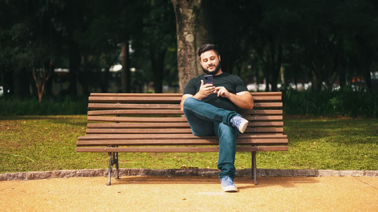 화창한 날 나무 공원 벤치에 혼자 앉아 웃고 있는 휴대폰을 사용하는 젊은 남자
