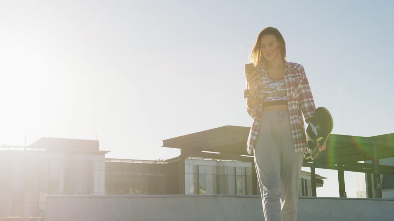 mujer caucásica, caminando, usando un teléfono inteligente y sosteniendo un skateboard en un skatepark