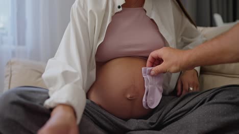 Close-up-a-man-attaches-small-baby-socks-to-his-pregnant-wife-belly-on-the-couch-at-home.-Husband-and-wife-are-expecting-a-child-and-preparing-clothes-for-his-birth