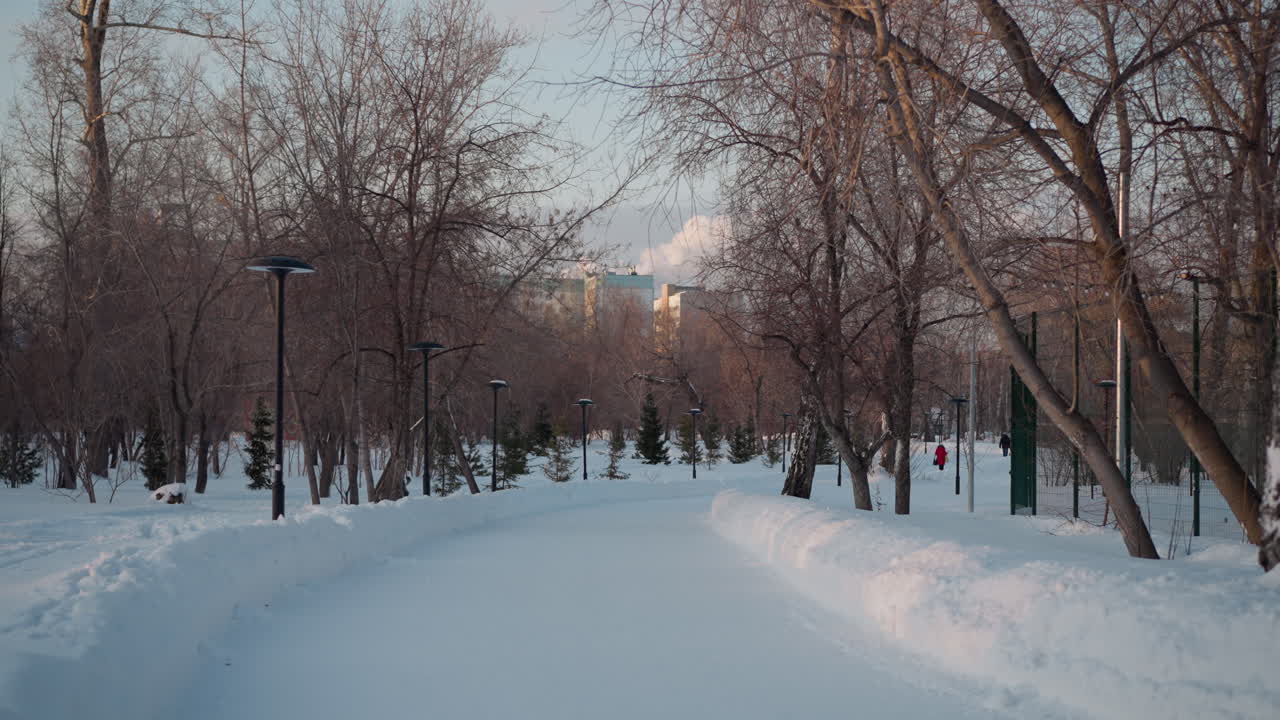 Winter season with person walking down snowy path lined with street lamps and bare trees, tall buildings in background, soft sunlight, peaceful winter city park atmosphere with shadows