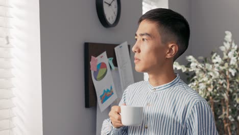 serious focused thoughtful head of the company, the businessman stands in the office of the corporate at the window covered with blinds and looks into it, contemplates the business