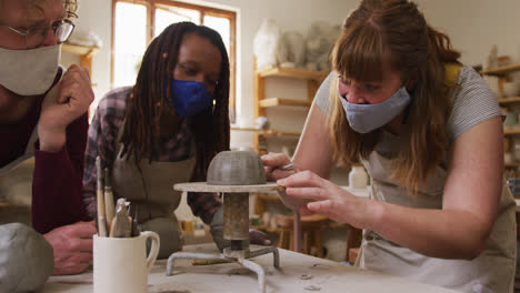 Diverse potters wearing face masks and aprons working on pot on potters wheel at pottery studio