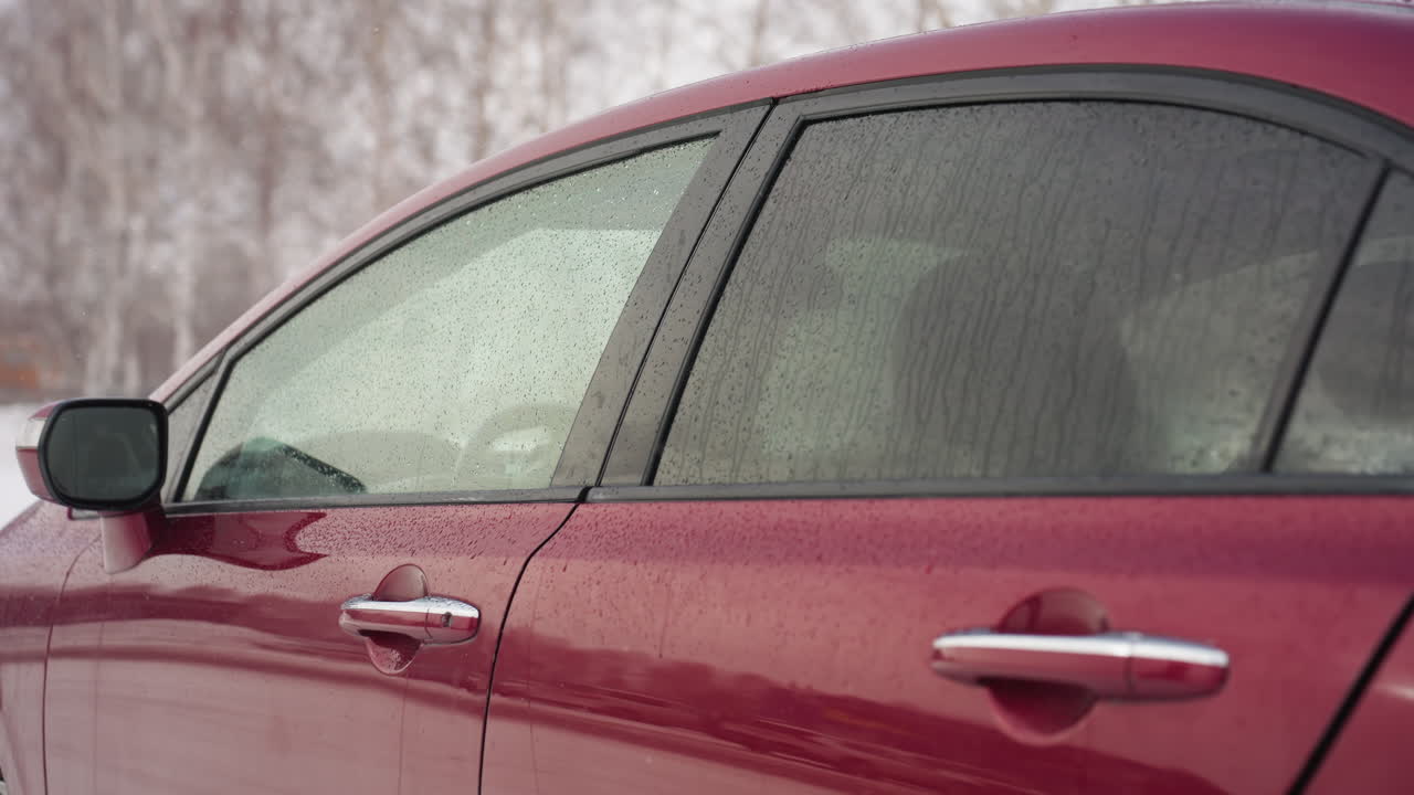 Boy in winter clothing closes red car door covered in water droplets from snowfall, with frosty surroundings and nearby structures reflecting on windows