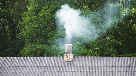 White-smoke-from-fireplace-rises-above-brick-chimney-on-residential-home-with-green-trees-in-background,-static