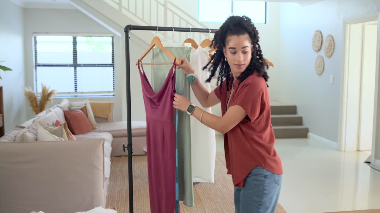 Mujer tomando una cinta métrica azul, midiendo un vestido malva en un perchero en casa, escribiendo tallas en un portátil