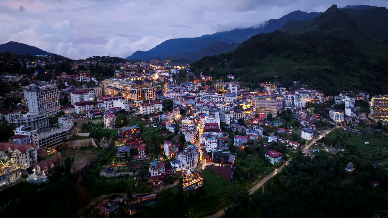 Establishing drone shot of Sapa town buildings, luxury hotels and resorts illuminated with night lights surrounded by mountains