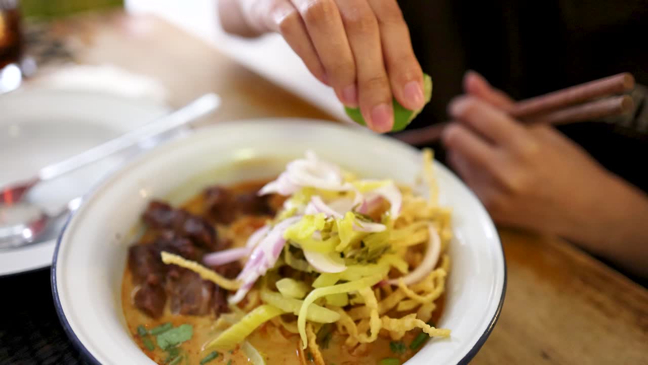 A hand squeezes fresh lime over a bowl of beef curry noodles with crispy toppings in a well-lit Bangkok restaurant, captured in a close-up sequence